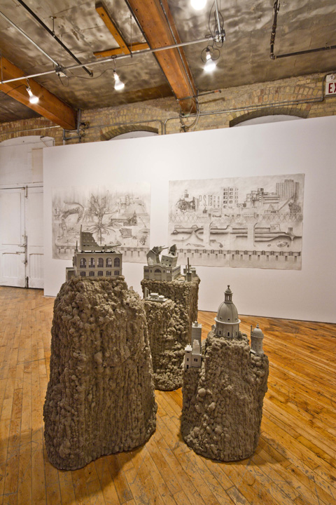 Grey building models sit on top of stalagmite like structures that rise from a wooden floor. Two large pencil and charcoal drawings hang in the background.