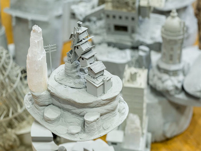 Betrayal at Babylon - detail of a larger sculpture that shows miniature kit bashed houses in a stack, sitting on a rocklike platform next to a tower shaped crystal.