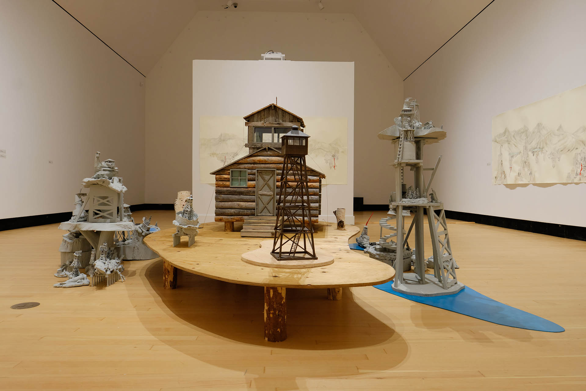 The Wilderness of Mirrors. Grouping of sculptures featuring fire tower models and a quarter scale log cabin arranged on a gallery floor.