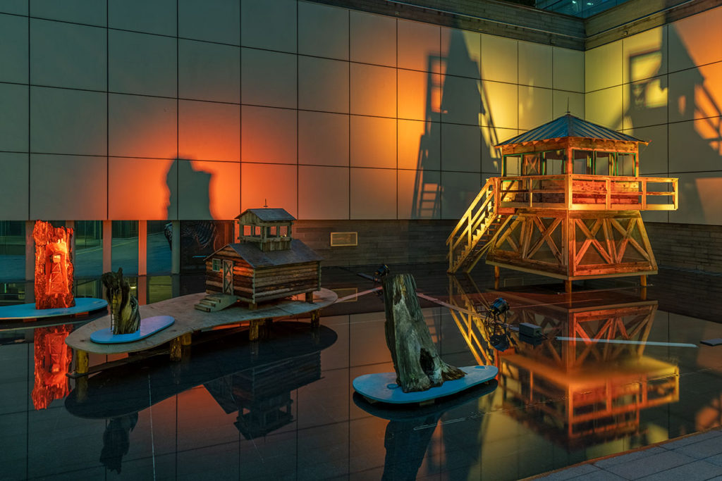 Grouping of sculptures in a flooded outdoor exihibition space made of dark concrete. From left to right sculptures include a stump with a small model firetower embedded, a quarter scale log cabin, a stump with a burnt hole at its base, a half scale two storey firetower.
