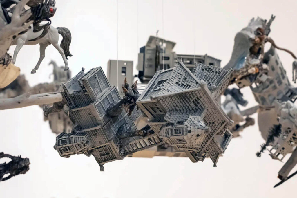 Photograph of a sculpture made of grey and black house and tower models, horses and ladders mashed together and suspended in the air.
