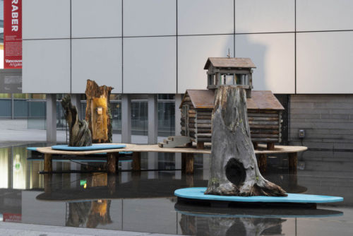 Grouping of sculptures in a flooded outdoor exihibition space made of dark concrete. From left to right sculptures include a stump with a small model firetower embedded, a quarter scale log cabin, a stump with a burnt hole at its base.