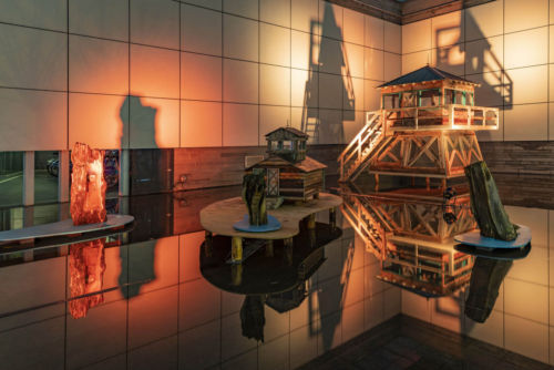 Grouping of sculptures in a flooded outdoor exihibition space made of dark concrete. From left to right sculptures include a stump with a small model firetower embedded, a quarter scale log cabin, a stump with a burnt hole at its base, a half scale two storey firetower.